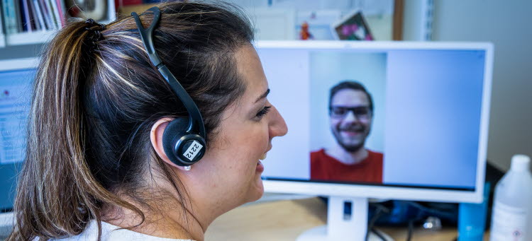 Kvinna med ett headset framför en skärm där en leende man syns.