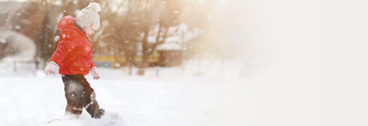 Ett barn som går och sparkar i snön, hen ler och har en röd jacka och mössa på sig.