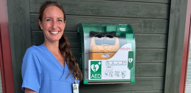 Frieda Jönsson från Lindsdals hälsocentral ler in kameran och står bredvid en hjärtstartare som hänger på väggen.