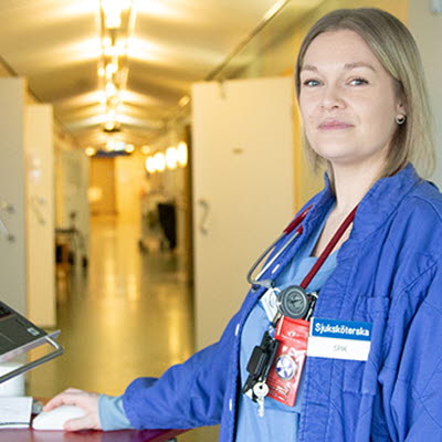 vårdklädd person står vid en dator i en sjukhuskorridor och tittar in i kameran