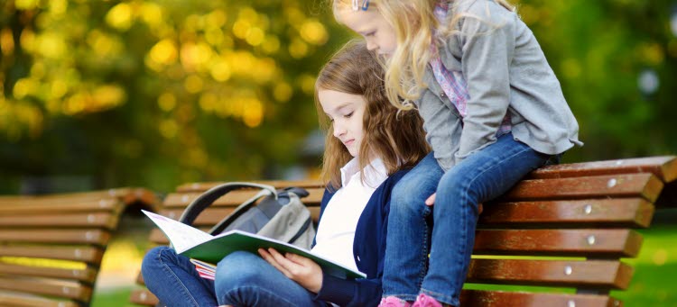 Två flickor sitter på en bänk och läser en bok