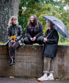 Tre unga kvinnor utomhus som ler mot varandra, en har ett svart paraply. 