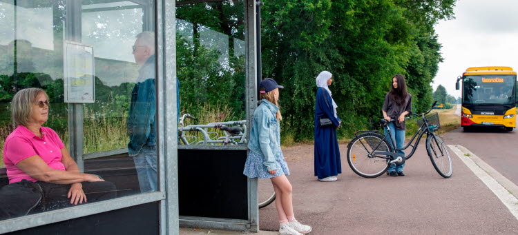 Buss svänger in på busshållplats där resenärer väntar.