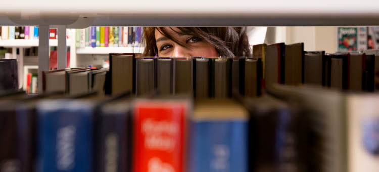 En kvinna tittar fram mellan böcker i en bokhylla. 