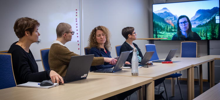 Fyra personer sitter runt ett konferensbord. En medarbetare är med på digital skärm.