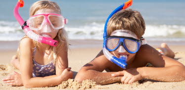 två barn ligger på en strand och har cyklop på sig, tittar in i kameran.