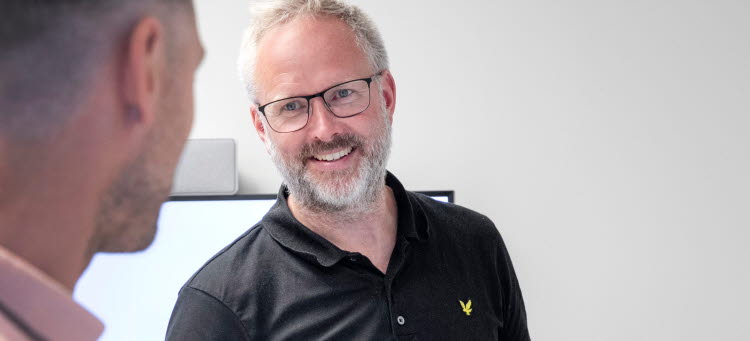 Torsten Dahlberg står med en laptop i handen och ler mot en kollega som står med ryggen mot kameran i förgrunden.
